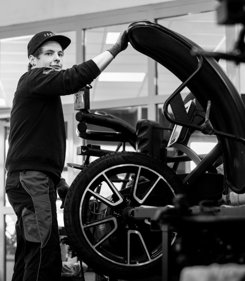 Ein junger Mitarbeiter arbeitet an einen Reifen in der Werkstatt und blickt lächelnd in die Kamera – weitere Reifen sind im Hintergrund zu sehen.