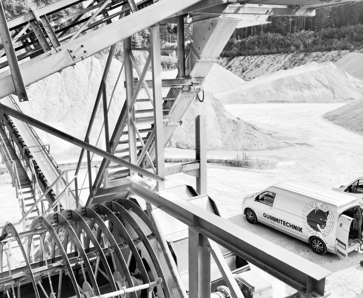 Übersicht einer großen Industrieanlage mit Silos, Förderbändern und Sand- oder Kieshügeln vor bewaldetem Hintergrund.