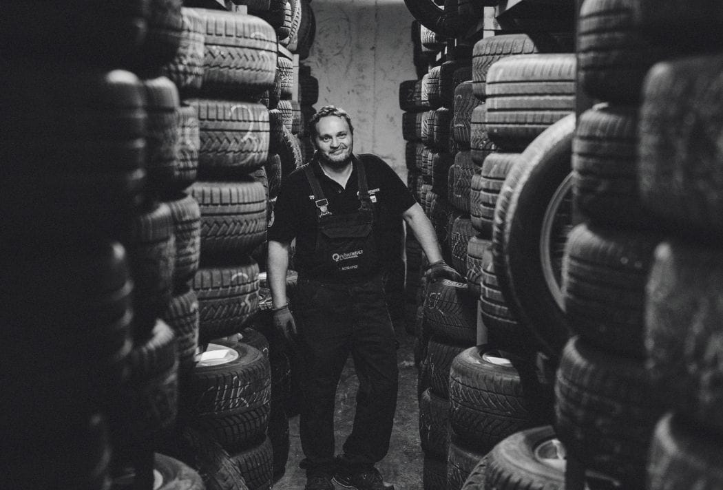 Ein Mitarbeiter steht in einem Lager voller großer Reifen und schaut konzentriert nach rechts – im Hintergrund sind Regale mit Reifen sichtbar.