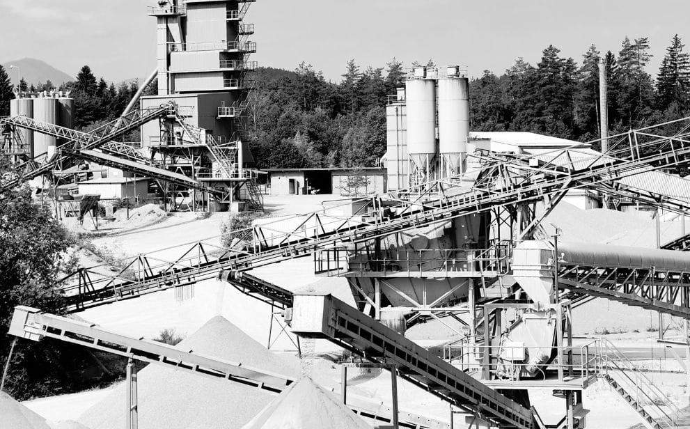 Übersicht einer großen Industrieanlage mit Silos, Förderbändern und Sand- oder Kieshügeln vor bewaldetem Hintergrund.